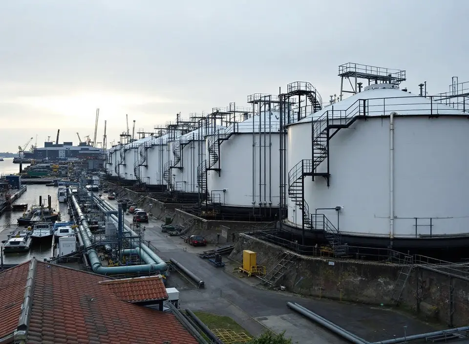 Een lange rij witte opslagtanks voor olie met wenteltrappen in een havengebied, met leidingen langs de kade en kranen in de verte